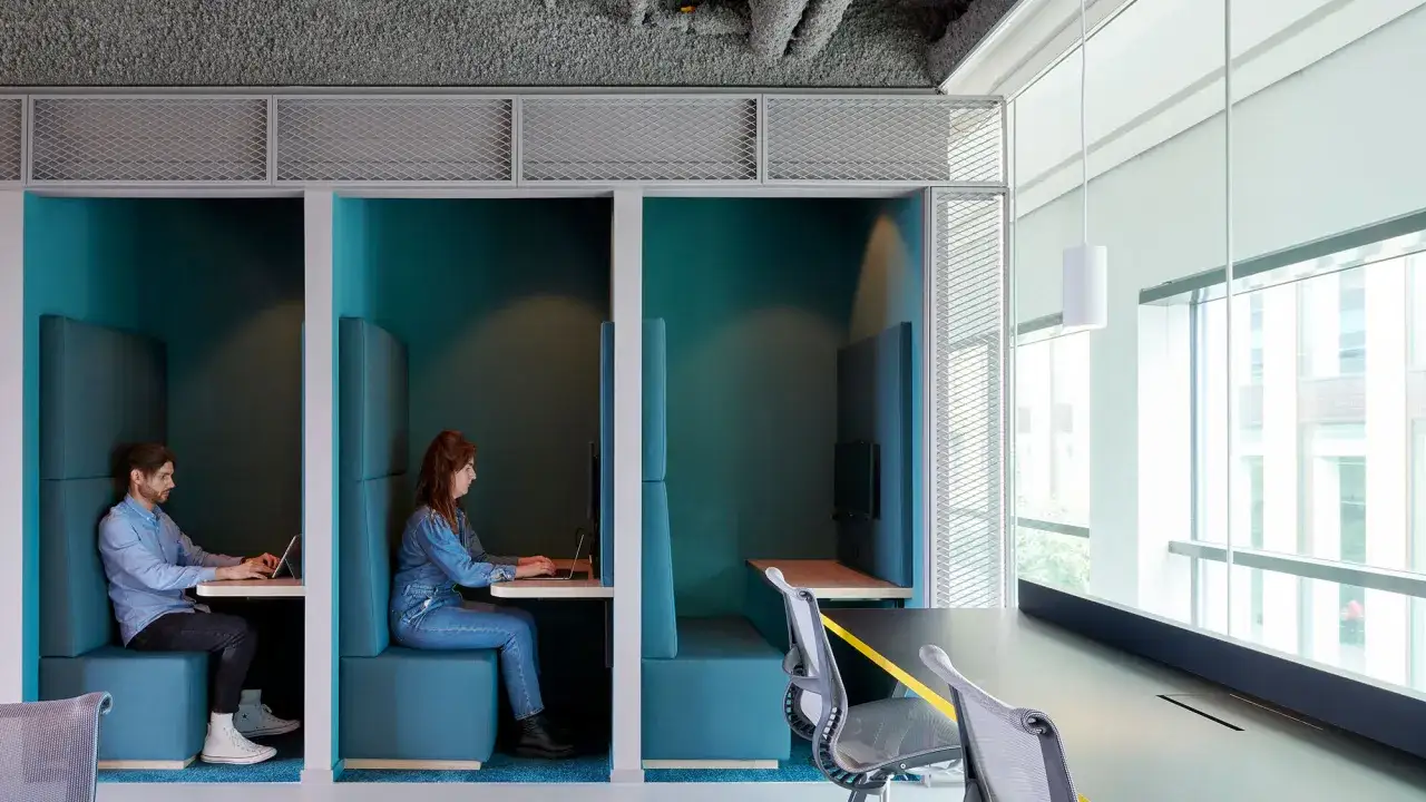 Two people work in separate, teal-colored pods within a modern open office space.
