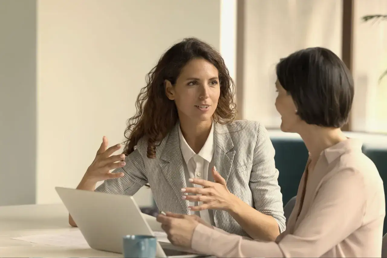 Two women discuss business, one explaining how to negotiate a raise with gestures.