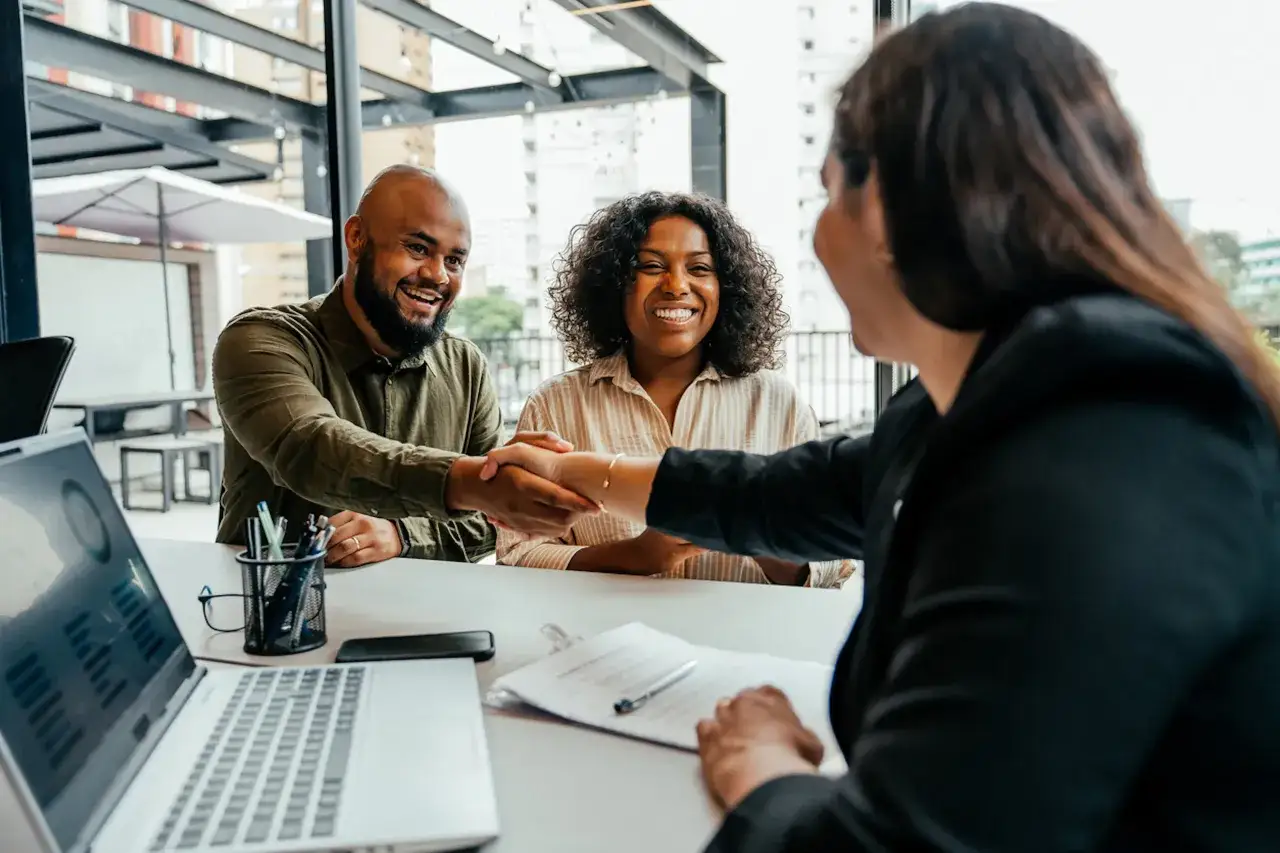 A man and woman smile as they shake hands, possibly concluding a positive employee feedback session. A laptop and documents are on the table.