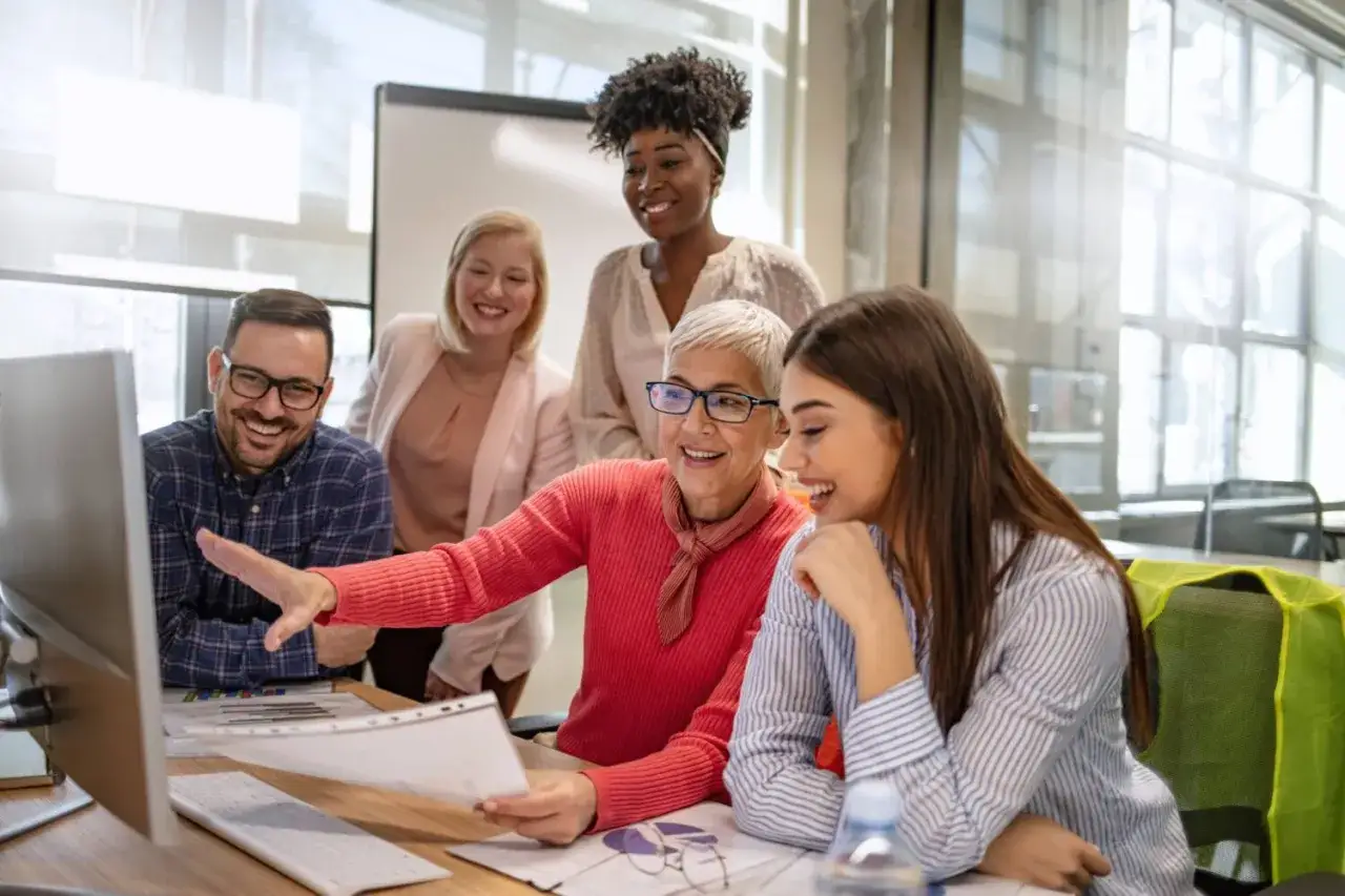 A diverse team collaborates, showcasing effective learning in the workplace as they review documents and a computer screen, all smiling.