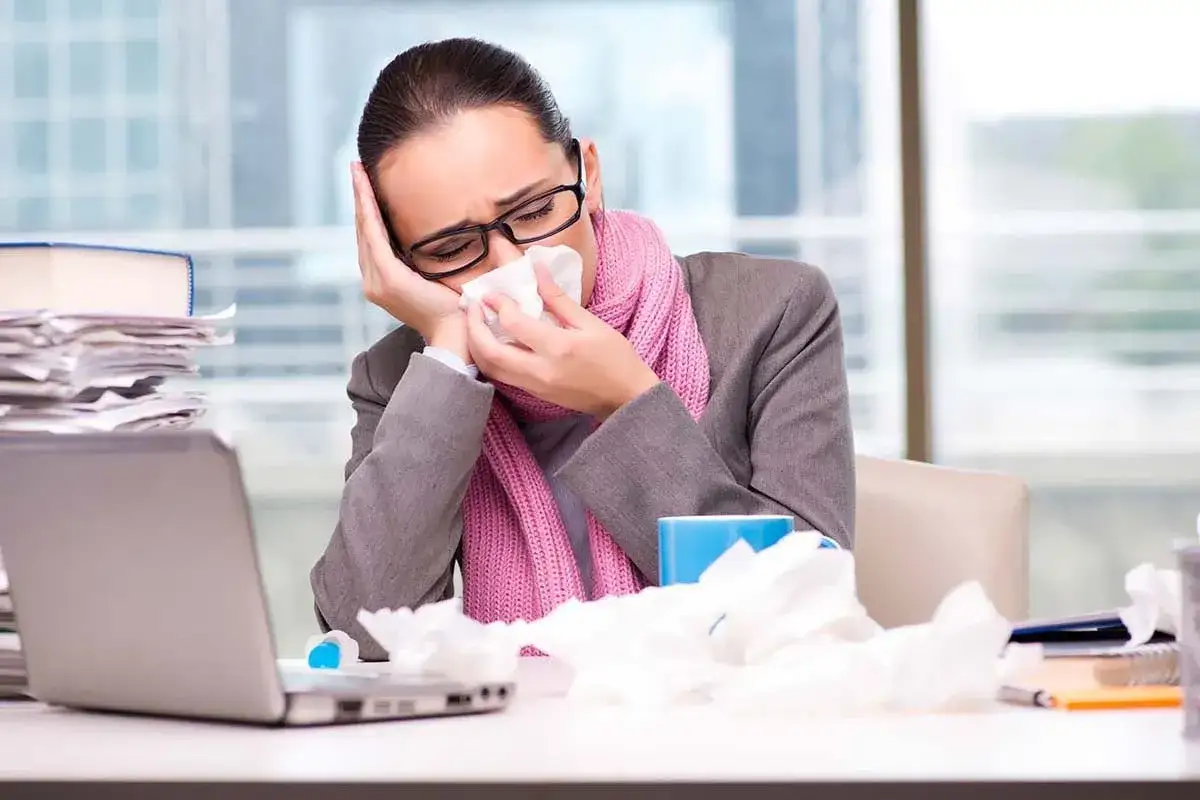 A sick woman with a pink scarf blows her nose into a tissue at her desk, surrounded by crumpled tissues and a laptop. This is presenteeism: working while unwell.