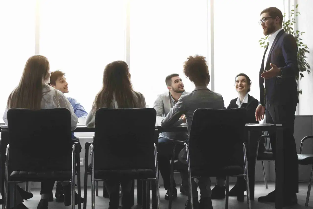 A man in a suit leads a CEO training and development session, gesturing to a group of engaged employees seated around a table.