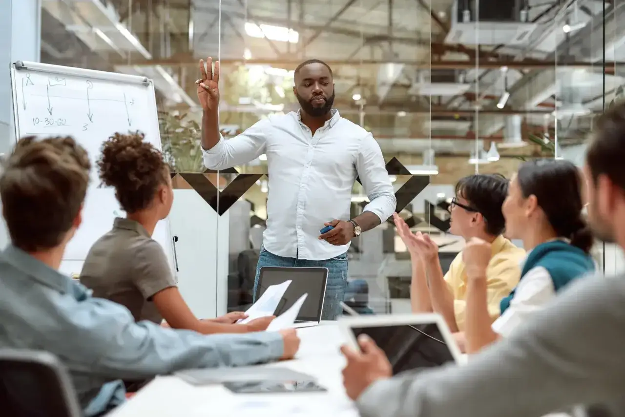 A trainer leads an empathy training session, gesturing with three fingers while a diverse group listens attentively.