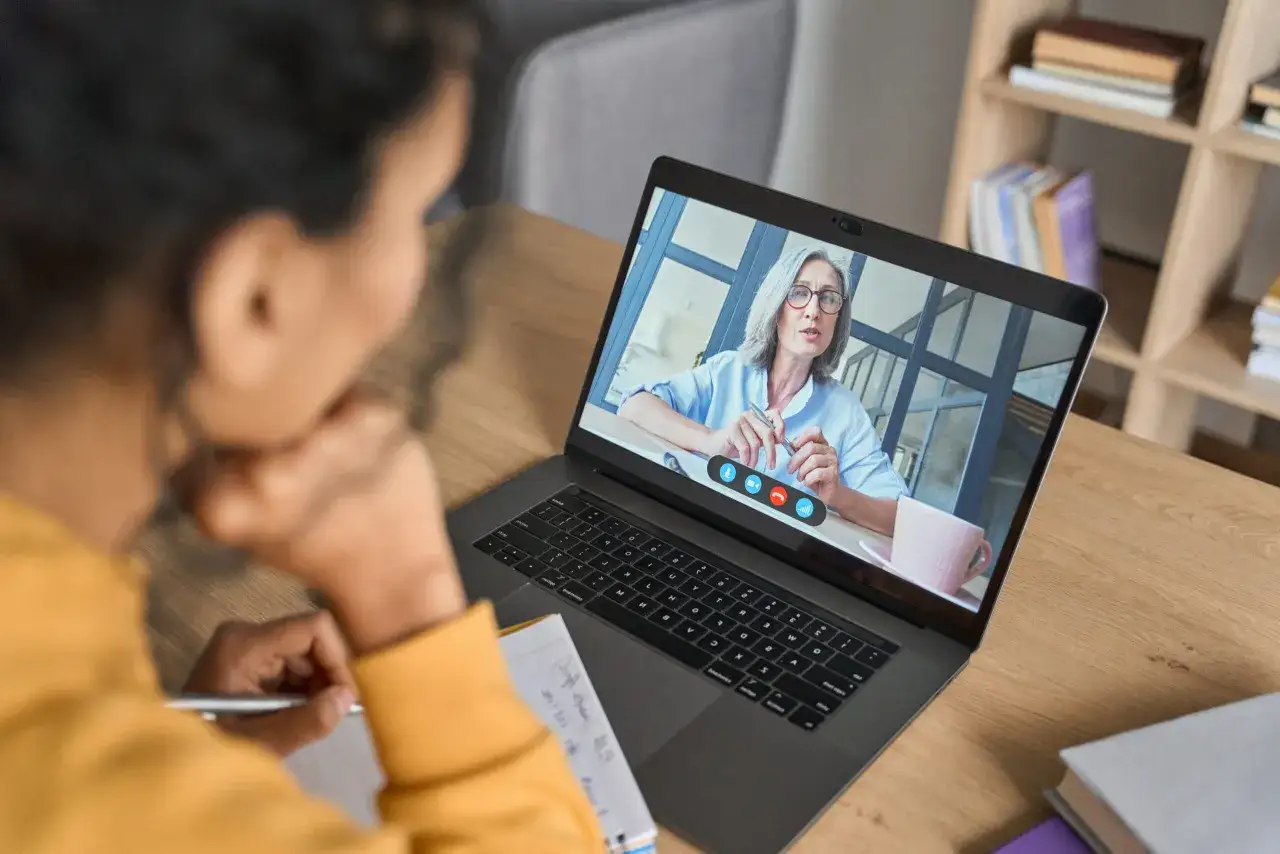 A woman takes notes during an online negotiation course. Her instructor appears on her laptop screen, offering guidance.