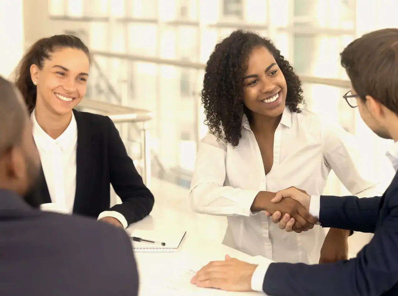 Two women and two men in a meeting. One woman shakes hands with a man, signifying a successful negotiation. A certificate of negotiation is likely being exchanged.