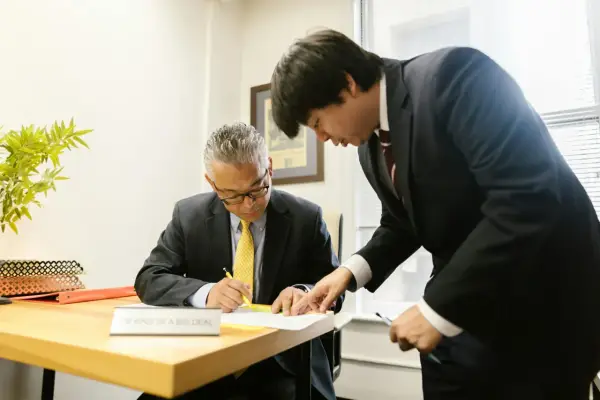 Two men in suits review documents. One man, seated, signs papers while the other, standing, points to the document. This scene prompts questions to ask your boss.
